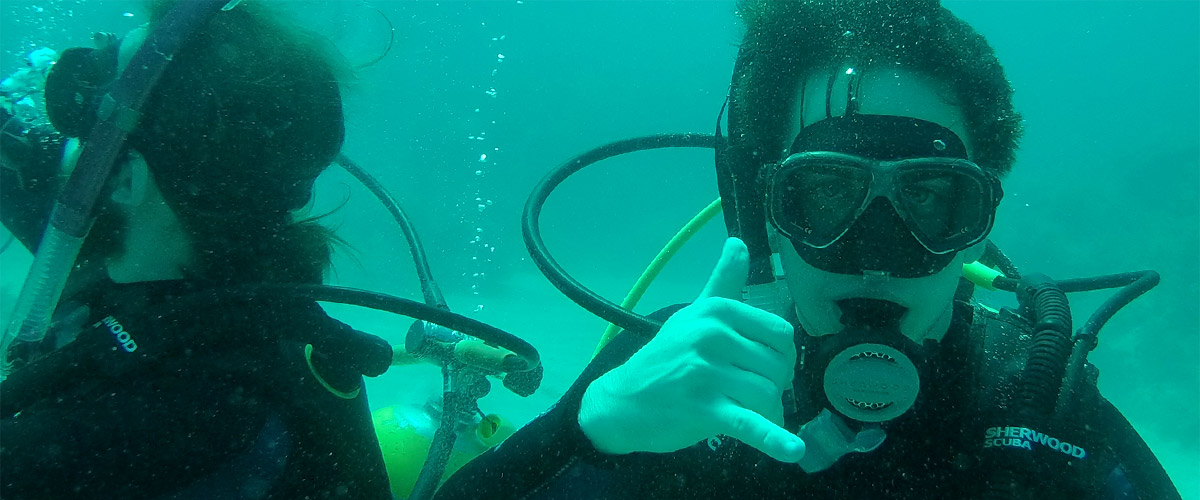 Image shows two students SCUBA diving. One is looking at the camera and making a “hang loose” (a.k.a. shaka) sign with her hand.