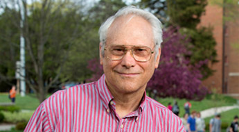 !A smiling Dr. Elton Graves standing outdoors on the Rose-Hulman campus with students walking in the background