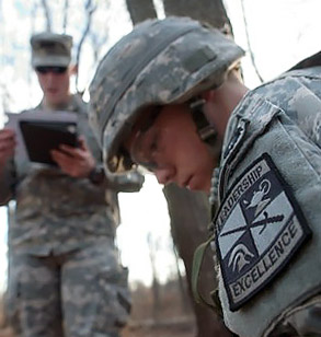 2 men dressed in uniform outside. One man is sitting and the other man is standing above him.