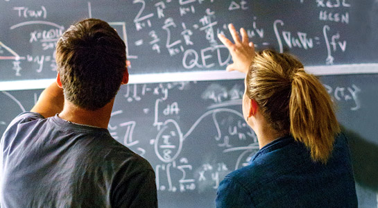 !Male and female students working a math problem on a chalkboard.