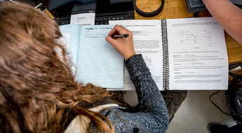 Female student solves physics equations in notebook.