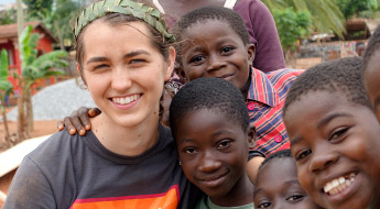 Female student wearing paper crown of leaves, hugging children in a village in Ghana. 