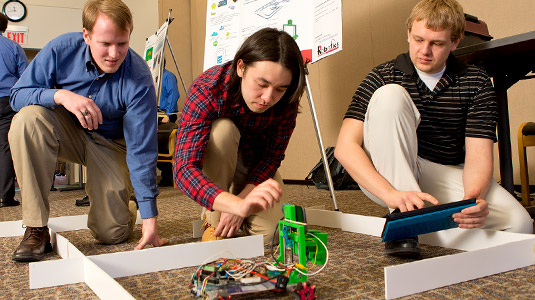 Students kneel to make adjustments to robot while using tablet to control device.