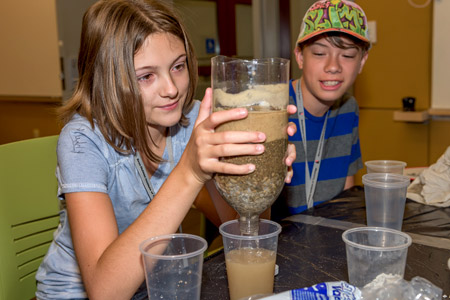 Female student holds upside-down two-liter bottle filled with layers of sand to make a water filter.