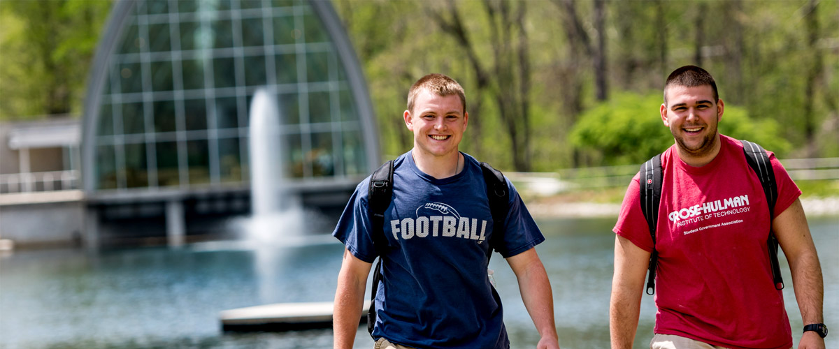 Two members of the Rose-Hulman football team smiling and walking along Speed Lake with White Chapel in the background.