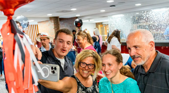 Parents lean in to take a selfie with a student at the Senior Celebration, surrounded by red streamers.