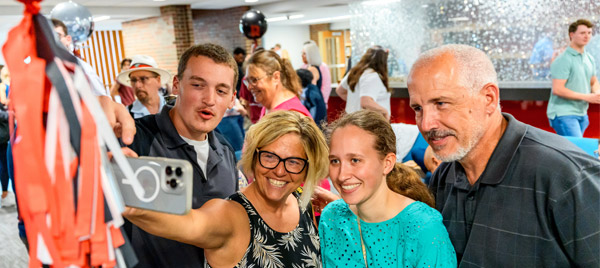 Parents lean in to take a selfie with a student during Senior Celebration, surrounded by red streamers.