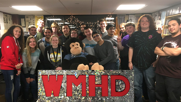 Staff of WMHD posing with a large stuffed gorilla. All are standing behind a large poster reading WMHD.