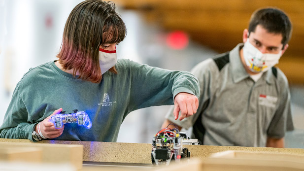 Image shows two students working with a small robot as it delivers pizza. 