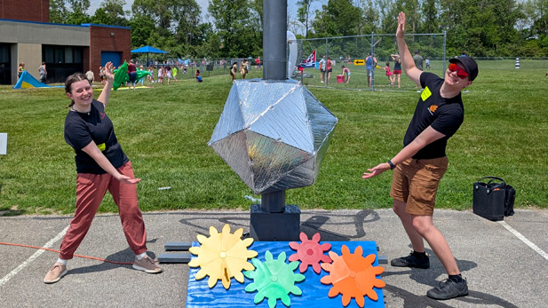 Two smiling volunteers pose enthusiastically beside a reflective geometric structure mounted on a pole, surrounded by colorful gear-shaped cutouts on the ground.