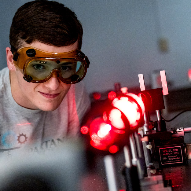 A student wearing protective orange laser safety glasses adjusts components in a laser optics lab setup.