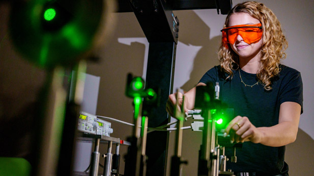 A student wearing protective orange laser safety glasses adjusts components in a laser optics lab setup.