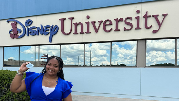 Jayden O'Dell in professional attire stands in front of Disney University holding her Walt Disney Imagineering name tag.
